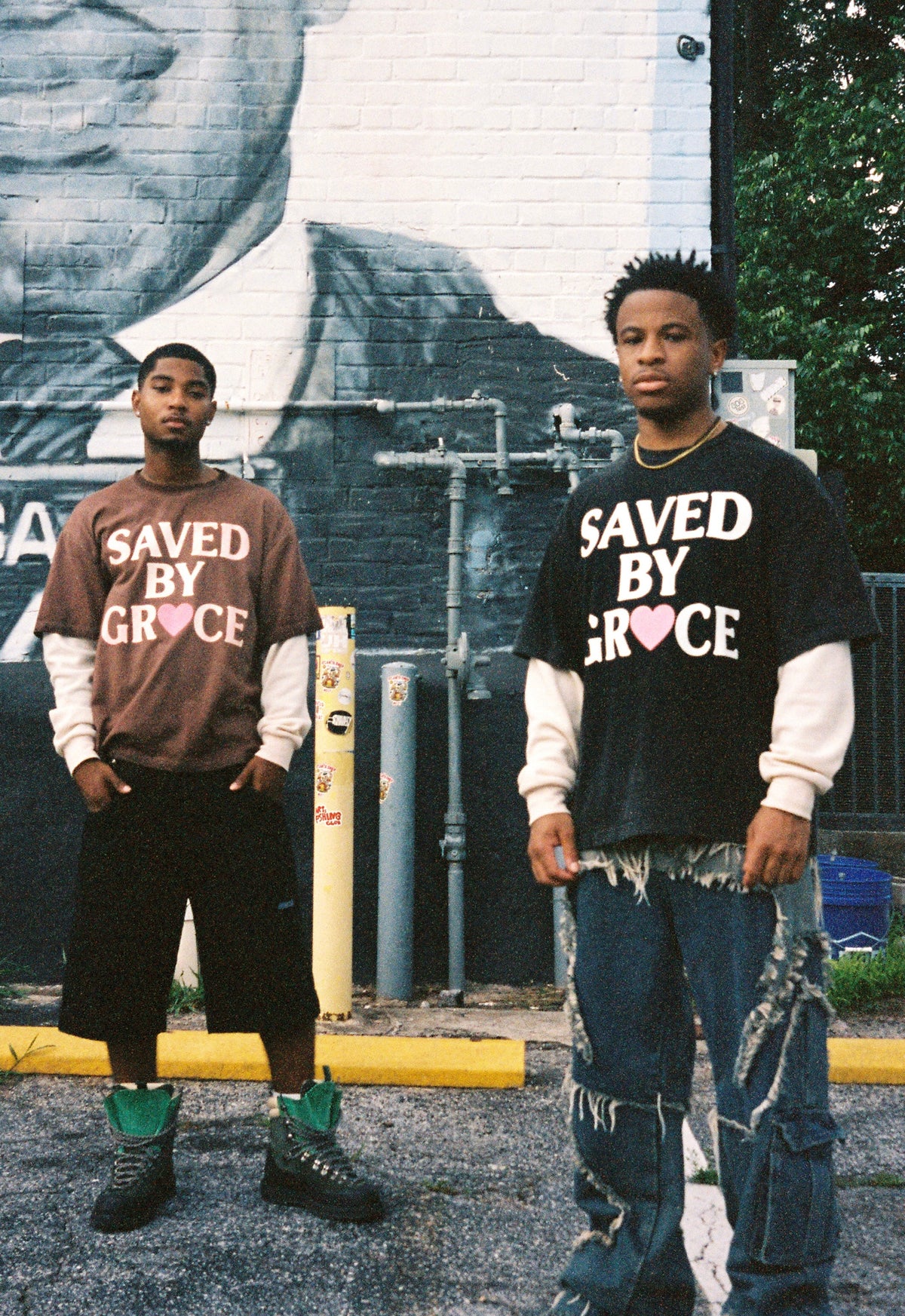 Two young men standing in front of a large black-and-white mural of Martin Luther King Jr. wearing graphic sweatshirts that read 'SAVED BY GRACE' and distressed jeans.